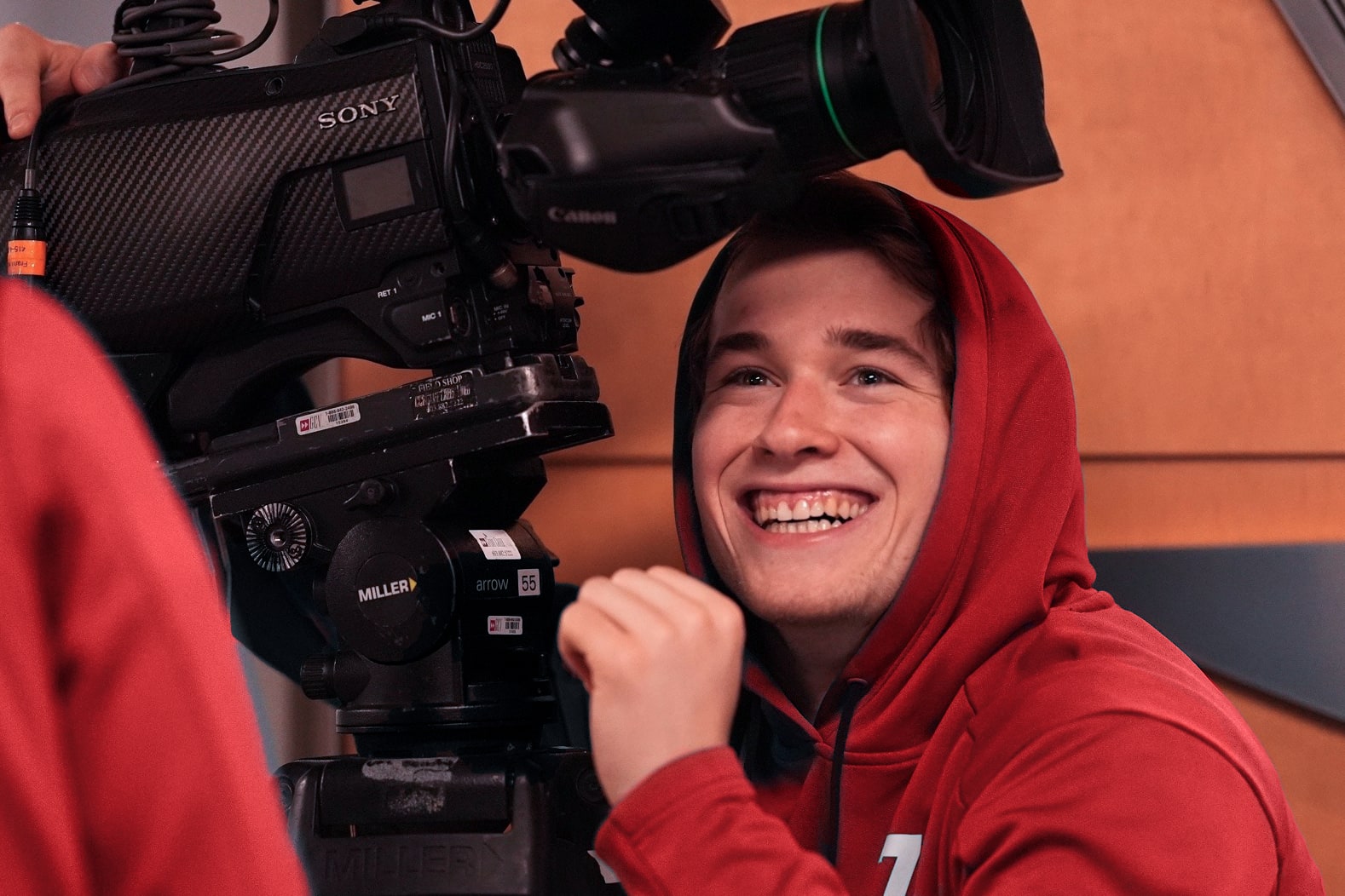 Luca Haas in a red hoodie hiding behind a camera rig
